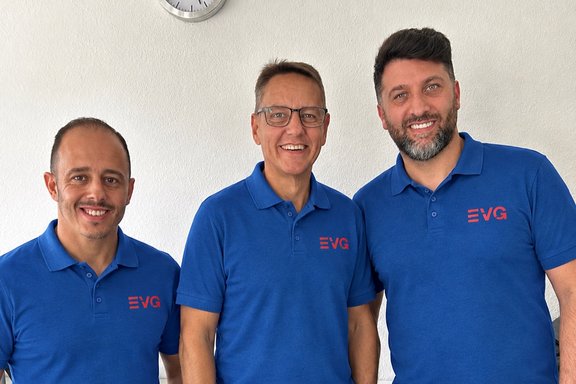 Drei Personen in blauen Poloshirts mit EVG-Logo stehen nebeneinander vor einer weißen Wand. Die drei Personen sind von der Hüfte aufwärts zu sehen und blicken in Richtung Kamera. Alle tragen gleichfarbige blaue Poloshirts. Auf der linken Brustseite der Shirts befindet sich das rote Logo der EVG (Eisenbahn- und Verkehrsgewerkschaft). Die Aufnahme wirkt wie ein Gruppen- oder Porträtfoto im Innenbereich.