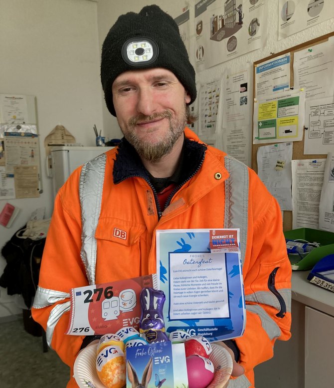 Eine Person in orangefarbener Warnschutzjacke mit reflektierenden Streifen und „DB“-Logo sowie schwarzer Mütze mit Stirnlampe steht in einem Innenraum. In den Händen hält sie einen geflochtenen Korb mit verschiedenen Gegenständen, darunter bunte Ostereier, Schokoladenhasen, eine Karte mit der Aufschrift „Ostergruß“, ein rotes Schild mit der Nummer „276“ und mehrere Artikel mit „EVG“-Logo. Im Hintergrund sind Pinnwände und Wände mit Aushängen, Plänen und Notizen zu sehen.