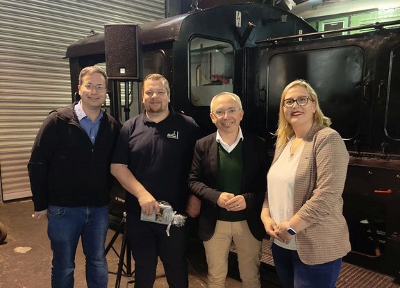 Vier Personen stehen nebeneinander in einer Halle vor einer schwarzen Lokomotive. Die Lokomotive ist seitlich zu sehen, mit Fenstern im Führerhaus und dunkler Metalloberfläche. Hinter den Personen befindet sich eine geriffelte Metallrolltür; links steht ein Lautsprecher auf einem Stativ. Die Personen tragen Freizeit- und Businesskleidung; eine Person hält einen kleinen, glänzend verpackten Gegenstand in den Händen. Der Boden ist dunkel, die Beleuchtung ist künstlich und gleichmäßig.