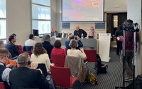 Innenraum eines hellen Konferenzraums mit Reihen roter Stühle. Im Vordergrund sitzen zahlreiche Personen, teils mit Notizblöcken, Laptops oder Smartphones; einige tragen Headsets. Vorn befindet sich ein Podium mit einem langen Tisch, hinter dem drei Personen sitzen. Auf dem Tisch stehen Namensschilder, Mikrofone und Wasserflaschen. Hinter dem Podium ist eine große Projektionsfläche mit einer Präsentationsfolie in Blau‑ und Orangetönen, mit Logos und mehrzeiligem Text, der aus der Entfernung nur teilweise lesbar ist. Rechts neben dem Podium steht ein Rednerpult mit Mikrofon. Am rechten Bildrand ist technisches Equipment zu sehen, darunter mehrere Kabel und ein Stativ. Der Raum hat hohe Fenster auf der linken Seite und einen gemusterten dunklen Teppichboden.