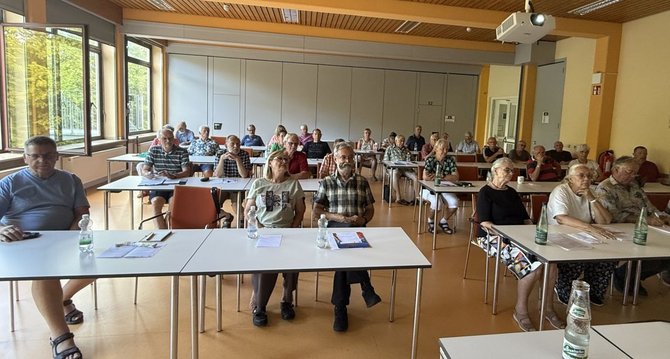 Seminarraum mit mehreren Tischen in Reihenanordnung. Rund 30 Personen sitzen verteilt an den Tischen und blicken nach vorn. Auf den Tischen stehen Wasserflaschen, Gläser, Notizblätter und Mappen. Links befinden sich große Fenster, an der Decke ein Beamer. Im Hintergrund sind Trennwände und Türen sichtbar.