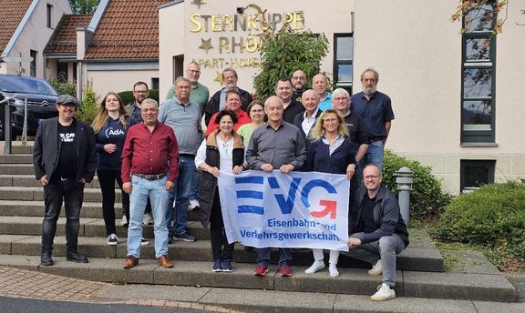 Etwa 16 Personen stehen auf einer Treppe vor einem Gebäude. In der vorderen Reihe halten mehrere Personen ein weißes Banner mit blau-rotem EVG-Logo und der Aufschrift „EVG Eisenbahn- und Verkehrsgewerkschaft“. An der Gebäudefassade steht groß „STERNKUPPE RHÖN APART-HOTEL“. Links ist ein geparktes Fahrzeug teilweise sichtbar, rechts befinden sich Fenster und Grünpflanzen. Die Personen tragen überwiegend Alltagskleidung und sind für ein Gruppenfoto angeordnet.