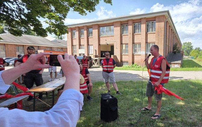 Mehrere Personen mit roten Warnwesten stehen oder sitzen auf einer Grünfläche vor einem Backsteingebäude. Rechts spricht eine Person in ein Handmikrofon, daneben steht ein Lautsprecher auf dem Boden. Im linken Vordergrund fotografiert eine Person die Szene mit einem Smartphone. Auf Tischen und Bänken liegen rote Fahnen.