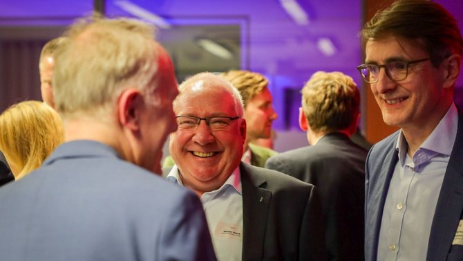 Drei Personen stehen in einem Innenraum nah beieinander. Die Person im Vordergrund links ist von hinten zu sehen und trägt ein Jackett. Die mittlere Person trägt eine Brille und ein Jackett mit Hemd und ist frontal sichtbar. Rechts steht eine weitere Person mit Brille, Hemd und Jackett. Im Hintergrund sind mehrere weitere Personen in einem beleuchteten Raum zu sehen.