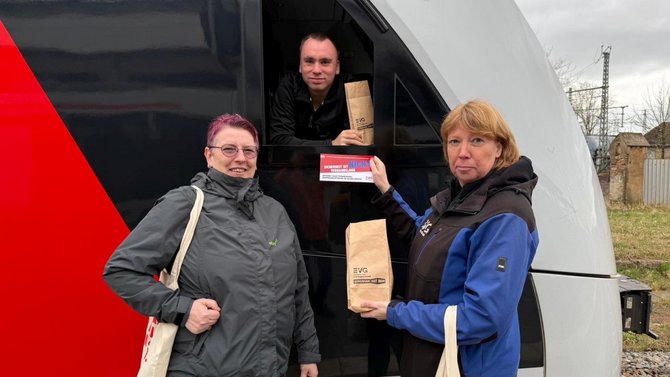 Eine Szene an einem Zug: Zwei Personen stehen außen vor einem modernen Zug und halten eine Papiertüte mit EVG‑Aufdruck in den Händen. Eine weitere Person hält aus dem geöffneten Fenster des Führerstands ebenfalls eine solche Tüte in den Händen. Eine der Personen draußen hält zusätzlich einen rechteckigen Infoflyer in der Hand. Im Hintergrund sind Gleisanlagen und einige Gebäude erkennbar.