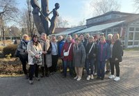 Eine Gruppe von etwa fünfzehn Personen steht dicht nebeneinander auf einem gepflasterten Weg im Freien. Die Personen tragen Jacken und Mäntel in verschiedenen Farben, einige mit Taschen oder Rucksäcken. Hinter der Gruppe befindet sich eine hohe Skulptur aus Holz, die zwei menschliche Figuren darstellt. Im Hintergrund sind ein Gebäude mit großen Fenstern und einem Glasdach sowie kahle Bäume zu sehen. Das Bild ist bei Tageslicht aufgenommen, der Himmel ist hell.