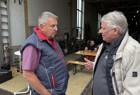 Zwei Personen stehen in einer Werkhalle oder an deren Eingang und unterhalten sich. Eine Person trägt eine blaue Steppweste über einem karierten Hemd, die andere eine helle Jacke über einem dunklen Oberteil. Im Hintergrund befinden sich Tische, Bänke, Gitterboxen und weitere Personen.