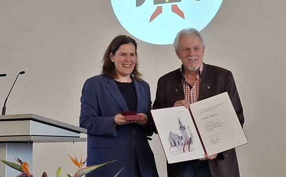 Zwei Personen stehen auf einer Bühne neben einem Rednerpult. Eine Person hält eine geöffnete Urkunde mit einer Kirchenillustration und sichtbarem Text, die andere hält ein kleines rotes Etui. Im Hintergrund ist ein projiziertes rundes Logo zu sehen. Vor der Bühne befinden sich Blumenarrangements mit orangefarbenen Blüten.