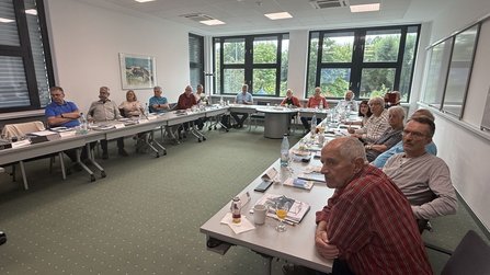 Heller Seminarraum mit U-förmig angeordneten Tischen. Rund 15 Personen sitzen an den Tischen. Auf den Tischen stehen Wasserflaschen, Gläser, Tassen, Unterlagen und Namensschilder. Große Fenster an der Rückseite geben den Blick auf Bäume frei.