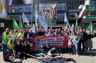 Große Gruppe von Menschen vor einem modernen Stadiongebäude mit Glasfassade. Im Hintergrund hängen Banner mit den gut lesbaren Texten „WIR ALLE FÜR WERDER“ und „WESERSTADION“. Mehrere Fahnen in Grün, Blau und Weiß werden hochgehalten, einige mit erkennbaren Gewerkschaftslogos. Im Vordergrund wird ein rotes Transparent gehalten mit dem Text „Erst unsere Jobs, dann eure Profite“ sowie dem DGB‑Logo. Weitere Fahnen und Banner liegen oder stehen vor der Gruppe.