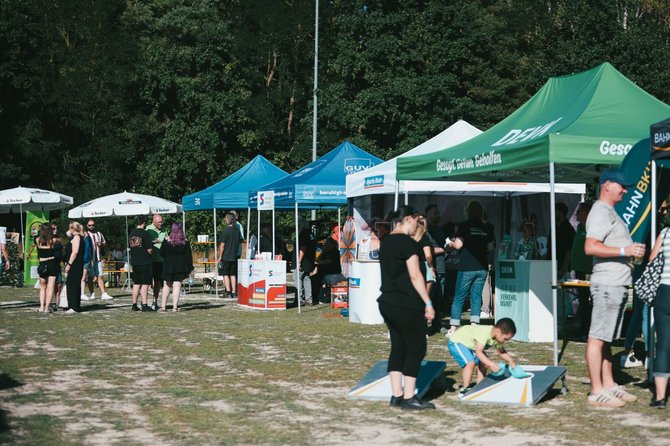 Mehrere Informations- und Aktionsstände mit Pavillons stehen auf einer freien Grasfläche vor einem bewaldeten Hintergrund. Zwischen den Ständen bewegen sich Besucherinnen und Besucher. Im Vordergrund spielt ein Kind an einem Wurfspiel, daneben geht eine Person über die Fläche. Auf den Pavillons sind verschiedene Schriftzüge und Logos sichtbar.