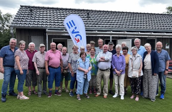 Gruppe von etwa 20 Personen auf einer Rasenfläche vor einem eingeschossigen Gebäude mit dunkler Holzfassade und Ziegeldach. In der Mitte steht eine weiße Fahne mit blau-rotem „IGR“-Schriftzug. Hinter der Gruppe sind geöffnete Türen und Fenster zu sehen.