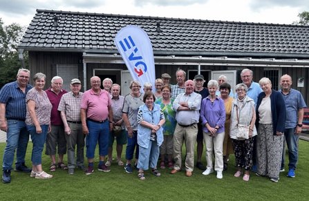 Gruppe von etwa 20 Personen auf einer Rasenfläche vor einem eingeschossigen Gebäude mit dunkler Holzfassade und Ziegeldach. In der Mitte steht eine weiße Fahne mit blau-rotem „IGR“-Schriftzug. Hinter der Gruppe sind geöffnete Türen und Fenster zu sehen.