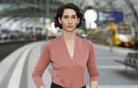 Person steht auf einem Bahnsteig eines großen Bahnhofs. Die Person trägt ein langärmeliges Oberteil in Rosé mit V-Ausschnitt, eine feine Halskette und kleine Ohrringe. Die Haare sind kurz und dunkel und leicht gewellt. Der Oberkörper ist frontal zur Kamera ausgerichtet, die Arme sind seitlich angewinkelt. Im Hintergrund sind Gleise, ein überdachter Bahnsteig mit Glasdach, unscharfe wartende Personen sowie ein gelbfarbener Zug zu sehen. Die Umgebung wirkt hell und weitläufig.