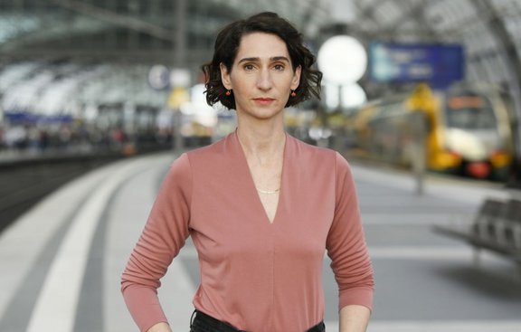 Person steht auf einem Bahnsteig eines großen Bahnhofs. Die Person trägt ein langärmeliges Oberteil in Rosé mit V-Ausschnitt, eine feine Halskette und kleine Ohrringe. Die Haare sind kurz und dunkel und leicht gewellt. Der Oberkörper ist frontal zur Kamera ausgerichtet, die Arme sind seitlich angewinkelt. Im Hintergrund sind Gleise, ein überdachter Bahnsteig mit Glasdach, unscharfe wartende Personen sowie ein gelbfarbener Zug zu sehen. Die Umgebung wirkt hell und weitläufig.