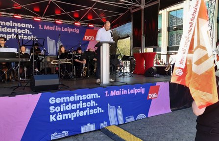 Bühne im Freien mit Rednerpult und Mikrofon. Eine Person steht am Pult, dahinter sitzt ein Musikensemble mit Instrumenten. An der Bühnenfront hängt ein großes Banner mit der Aufschrift „Gemeinsam. Solidarisch. Kämpferisch. 1. Mai in Leipzig“ sowie dem Logo „DGB“. Rechts ist eine orange Fahne teilweise im Bild zu sehen.