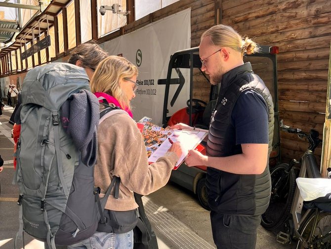 Drei Personen stehen in einem überdachten Bahnhofsbereich dicht beieinander. Eine Person mit Rucksack beugt sich leicht nach vorn, eine zweite Person hält ein Klemmbrett mit Papieren, eine dritte Person zeigt mit einem Stift auf das Dokument. Im Hintergrund sind eine Holzwand, ein kleines Fahrzeug und abgestellte Fahrräder sichtbar.