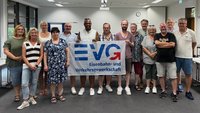 Eine Gruppe von Personen steht in einem Seminarraum zusammen und hält ein Banner mit der Aufschrift „EVG Eisenbahn- und Verkehrsgewerkschaft“. Insgesamt stehen etwa 14 Personen nebeneinander in einer Reihe und blicken zur Kamera. In der Mitte halten mehrere Personen ein großes, weißes Banner, auf dem in blauer Schrift „EVG“ sowie darunter „Eisenbahn- und Verkehrsgewerkschaft“ zu lesen ist. Die Personen tragen Alltagskleidung wie T‑Shirts, Hemden, Kleider und kurze Hosen.
