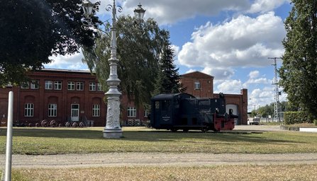 Freifläche vor einem roten Backsteingebäude mit weißen Fenstern. Im Vordergrund steht ein historisch wirkender, weiß verzierter Laternenmast auf einer Rasenfläche. Daneben befindet sich ein kleines dunkles Schienenfahrzeug auf einem kurzen Gleisabschnitt. Im Hintergrund sind Bäume, weitere Gebäudeteile und ein teilweise bewölkter Himmel sichtbar.