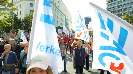 Demonstrationszug auf einer breiten Straße. Mehrere weiße Fahnen mit blauem und rotem Aufdruck sind im Vordergrund sichtbar. Zahlreiche Teilnehmende gehen zwischen Gebäuden und Bäumen die Straße entlang.