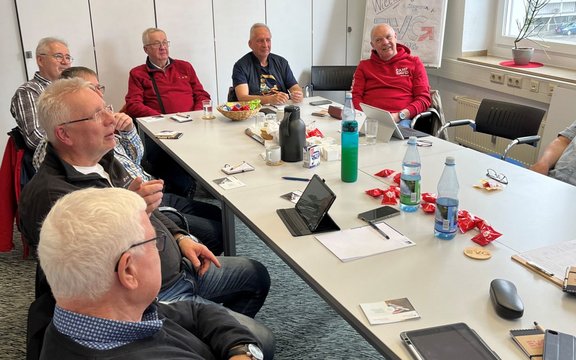 Eine Gruppe von Personen sitzt um einen rechteckigen Konferenztisch in einem Besprechungsraum. Auf dem Tisch stehen Getränke, Kaffeetassen, Snacks, Tablets, Notizblöcke und Schreibmaterial. Im Hintergrund sind eine Flipchart, Schränke und ein Fenster mit einer Topfpflanze sichtbar.