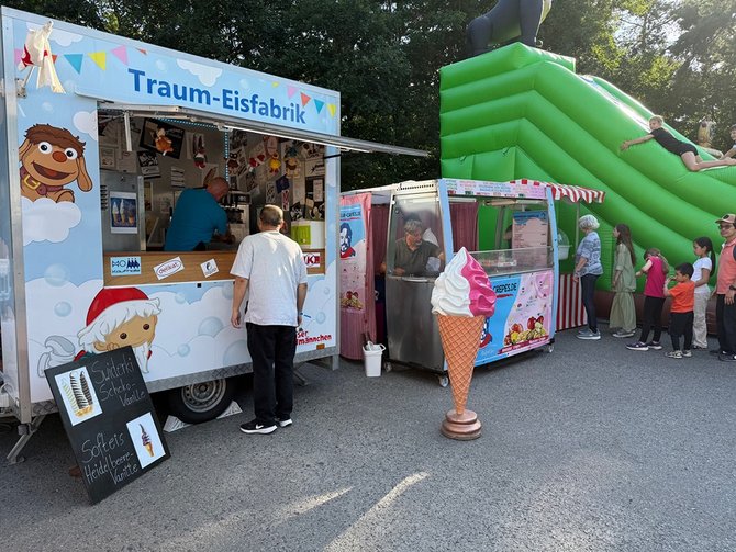 Ein Eisstand und eine Hüpfburg stehen bei einem Fest im Freien, während mehrere Menschen anstehen oder sich anstellen. Links im Bild steht ein Verkaufswagen mit der Aufschrift „Traum‑Eisfabrik“. Vor dem Wagen stehen zwei Personen an der Ausgabe. Neben dem Wagen steht eine kleine Tafel mit den Angeboten, zum Beispiel Softeis und andere Eissorten. Rechts daneben befindet sich ein weiterer kleiner Stand sowie eine große dekorative Eiswaffel aus Kunststoff. Im rechten Bereich des Bildes steht eine große, grüne Hüpfburg mit Rutsche. Ein Kind rutscht gerade herunter, während mehrere weitere Kinder und Erwachsene daneben in einer Reihe warten. Die Szene spielt im Freien auf einer befestigten Fläche, umgeben von Bäumen. Das Wetter wirkt sonnig und hell.