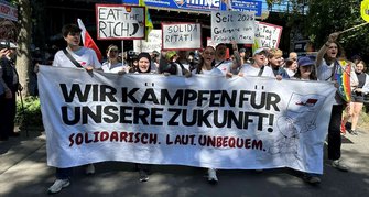 Demonstrationszug auf einer Straße mit Bäumen am Rand. Mehrere Personen halten ein großes weißes Banner mit der Aufschrift: „WIR KÄMPFEN FÜR UNSERE ZUKUNFT! Solidarisch. Laut. Unbequem.“ Dahinter sind weitere Plakate zu sehen, darunter „EAT THE RICH“, „SOLIDARITÄT!“ und „Seit 2025 Gefangene von Friedrich Merz“. Fahnen und weitere Schilder sind im Hintergrund erkennbar.