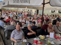 Große Gruppe von Menschen sitzt dicht gedrängt an Tischen in einem Außenbereich einer Gaststätte unter mehreren Sonnenschirmen mit der Aufschrift „Tucher – Bier aus Nürnberg“. Die meisten Personen sind älter, sitzen auf Stühlen um Tische mit Getränkegläsern, Tassen und Flaschen. Im Hintergrund sind Hausfassaden mit Fenstern und Blumenkästen zu sehen.