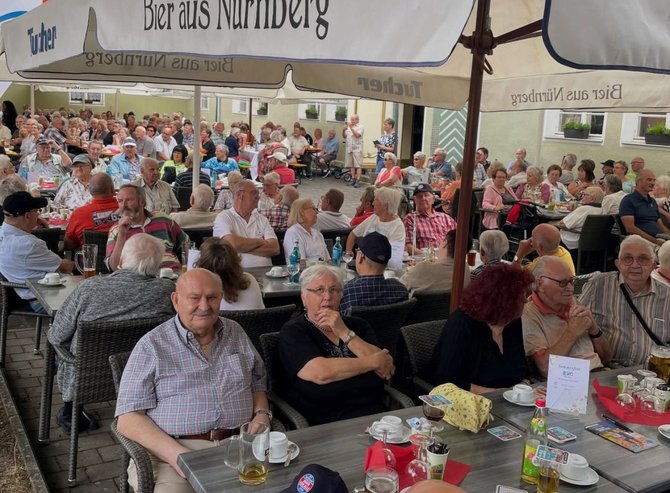 Große Gruppe von Menschen sitzt dicht gedrängt an Tischen in einem Außenbereich einer Gaststätte unter mehreren Sonnenschirmen mit der Aufschrift „Tucher – Bier aus Nürnberg“. Die meisten Personen sind älter, sitzen auf Stühlen um Tische mit Getränkegläsern, Tassen und Flaschen. Im Hintergrund sind Hausfassaden mit Fenstern und Blumenkästen zu sehen.