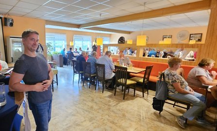 Gastraum mit hellen Wänden, mehreren langen Tischen und Stühlen. Im linken Vordergrund steht eine Person neben einem Stehtisch. An den Tischen sitzen zahlreiche Personen. Über den Sitzbereichen hängen rechteckige Leuchten, an der Wand befinden sich Spiegel, Fenster und Holzverkleidungen.