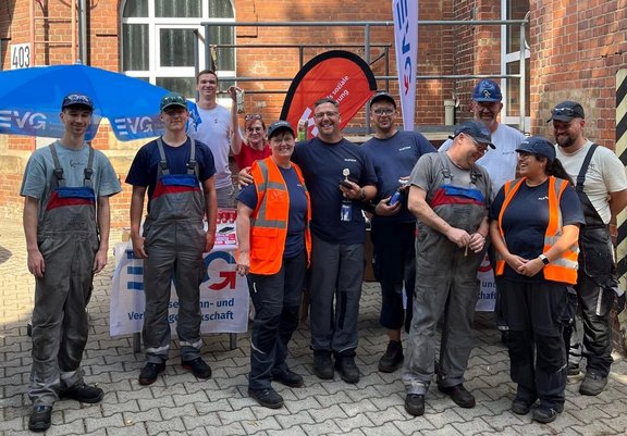 Gruppenfoto im Freien vor einem Backsteingebäude: Zehn Personen stehen vor zwei Tischen mit EVG-Bannern und blauen Sonnenschirmen. Mehrere Personen tragen graue Arbeitskleidung, einige zusätzlich orangefarbene Warnwesten oder Schutzhelme. Hinter der Gruppe sind Fahnen und Werbematerialien zu sehen. Auf dem Gebäude ist die Hausnummer „403“ sichtbar. Die Aufnahme zeigt die Gruppe von Kopf bis Fuß auf einem gepflasterten Hof.