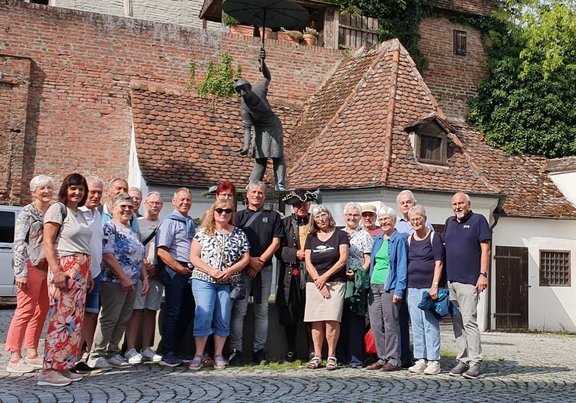 Etwa 16 Personen stehen auf einem gepflasterten Platz vor einer bronzefarbenen Statue, die eine Person mit erhobenem Schirm darstellt. Hinter der Gruppe befinden sich Gebäude mit roten Ziegeldächern, weißen Fassaden und einer hohen Backsteinmauer. Rechts ist eine mit Efeu bewachsene Wand zu sehen. Das Bild zeigt eine sonnige Außenaufnahme.