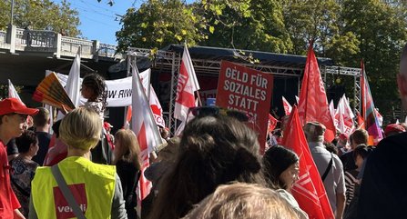 Menschenmenge vor einer Bühne im Freien. Mehrere rote und weiße Fahnen ragen über die Menge. In der Bildmitte ist ein Schild mit der Aufschrift „GELD FÜR SOZIALES STATT“ zu sehen. Links trägt eine Person eine gelbe Weste mit ver.di-Logo. Bäume und eine Brücke befinden sich im Hintergrund.