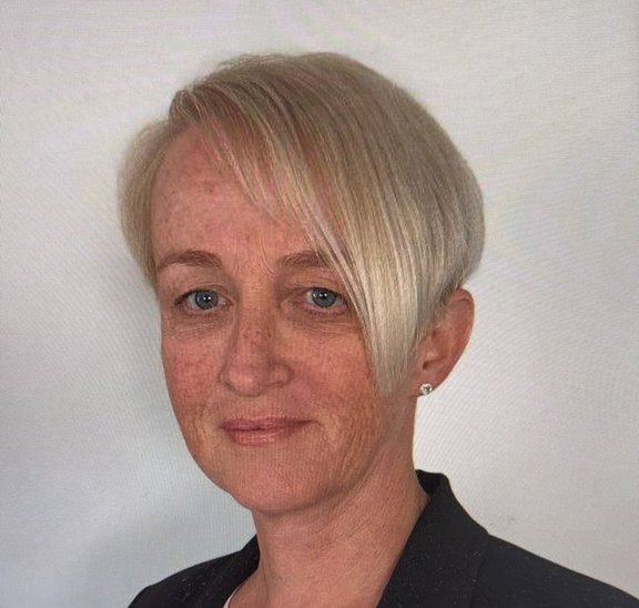 Porträt einer Person mit kurzem, hellblondem Haar vor einem hellen, einfarbigen Hintergrund. Die Person trägt ein dunkles Oberteil und kleine Ohrstecker und blickt in die Kamera.