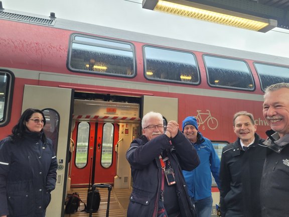 Gruppe von Menschen am Bahnhof, die vor einem roten Zug stehen und fröhlich miteinander interagieren, während es leicht regnet.