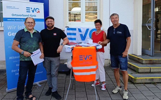 Vier Personen stehen vor einem Gebäude neben einem Infostand mit einem Banner „EVG Ortsverband Fürth“ und einer gut sichtbaren orangefarbenen Warnweste mit Logo am Tisch, auf dem Informationsmaterialien ausliegen.