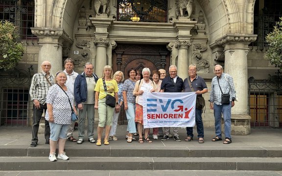 Gruppe von etwa 13 älteren Menschen steht auf einer Treppe vor einem reich verzierten steinernen Eingang mit Säulen und Bögen. In der Mitte halten mehrere Personen ein weißes Banner mit der Aufschrift „EVG SENIOREN“ und kleinerem Text darunter. Die Personen sind unterschiedlich gekleidet und schauen überwiegend in die Kamera.
