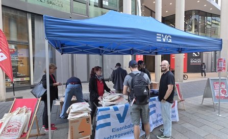 Blauer Pavillon mit dem Aufdruck „EVG“ über einem Informationsstand. Auf dem Tresen liegen Stapel von Stofftaschen und Informationsmaterial. Mehrere Personen stehen am Stand oder bewegen sich darum herum. Links und rechts sind rote Liegestühle und Werbeaufsteller zu sehen. Im Hintergrund befinden sich moderne Gebäude mit großen Glasflächen.