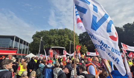 Menschenmenge auf einer Kundgebung im Freien vor einer Bühne. Mehrere Fahnen und Transparente werden über der Menge gehalten, darunter weiße Fahnen mit blauem und rotem Aufdruck sowie rote Fahnen. Links steht ein großes Gebäude mit Glasfassade, im Hintergrund sind Bäume zu sehen. Der Himmel ist teilweise bewölkt.