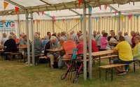 Großes Festzelt mit langen Bierbänken und Tischen auf einer Rasenfläche. Zahlreiche Personen sitzen in Gruppen an den Tischen. Über den Sitzreihen hängen bunte Wimpelketten. Links ist ein Banner mit der Aufschrift „EVG Eisenbahn- und Verkehrsgewerkschaft“ zu sehen. Im Vordergrund steht eine rote Gehhilfe neben einer Bank.
