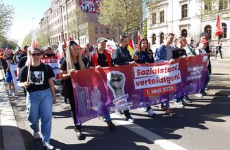 Eine größere Gruppe von Menschen geht auf einer Straße und hält ein breites Banner. Auf dem Banner stehen die gut lesbaren Texte „Sozialstaat verteidigen!“, „1. Mai 2026“, „Erst unsere Jobs, dann eure Profite“ sowie „Rente, Miete und Pflege nicht verkehrt“. Mehrere Fahnen werden mitgetragen. Die Straße ist von Gebäuden gesäumt, Bäume mit frischem Laub stehen am Rand, und weiteres Publikum befindet sich im Hintergrund.
