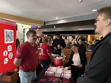 Informationsstand mit ausgelegten weißen Textilien und Schlüsselbändern auf einem Tisch. Links steht ein roter Aufsteller mit QR-Code und Symbolen für soziale Medien. Mehrere Personen stehen am Tisch, halten Informationsmaterial oder Stofftaschen in den Händen. Im Hintergrund befindet sich ein Bereich mit der Beschriftung „ABHOLSTATION“, Verkaufsautomaten und weitere Besucherinnen und Besucher.