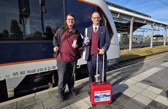 Person mit Brille und dunkler Jacke steht in einem Zug oder Bahnfahrzeug. Auf der Jacke ist ein kleines „DB“-Logo sichtbar. Die Person trägt einen Rucksack und hält in einer Hand eine kleine transparente Tüte mit roten und weißen Elementen, in der anderen Hand mehrere längliche Gegenstände. Im Hintergrund sind ein Fenster mit Blick auf Gleise und Vegetation sowie ein Bildschirm zu sehen.