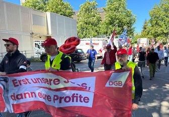 Straßenumzug mit vielen Personen auf einer gepflasterten Straße. Im Vordergrund tragen mehrere Personen Warnwesten und halten ein großes rotes Banner mit der Aufschrift „Erst unsere Löhne, dann eure Profite“ und dem Logo „DGB“. Dahinter folgen weitere Personen mit Fahnen und Schildern, darunter EVG-Fahnen. Am Straßenrand stehen Gebäude, Bäume und geparkte Fahrzeuge. Tageslicht.