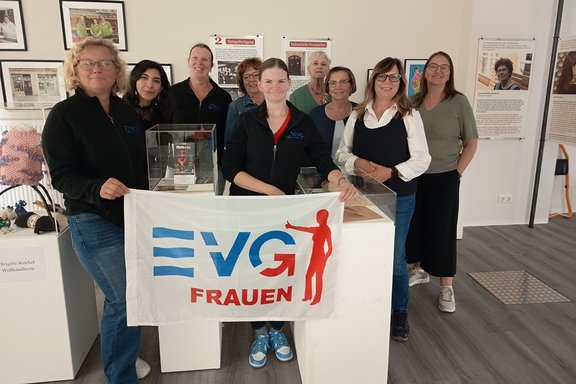 Eine Gruppe von Personen steht in einem Ausstellungsraum hinter einer Fahne mit der Aufschrift „EVG FRAUEN“ und posiert für ein Gruppenfoto. Im Vordergrund halten zwei Personen eine weiße Fahne mit dem blauen und roten EVG-Logo sowie dem Schriftzug „FRAUEN“. Die Gruppe umfasst mehrere Personen, die sich hinter und neben der Fahne aufgestellt haben. Einige tragen dunkle Jacken mit EVG-Logo.