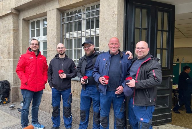 Fünf Personen stehen nebeneinander vor einer steinernen Gebäudefassade mit Fenstern und einer offenen Tür. Die Personen tragen Arbeitskleidung und Jacken und halten rote Becher in den Händen. Der Boden ist gepflastert; durch die geöffnete Tür ist ein Innenraum mit Tischen und weiteren Personen erkennbar.