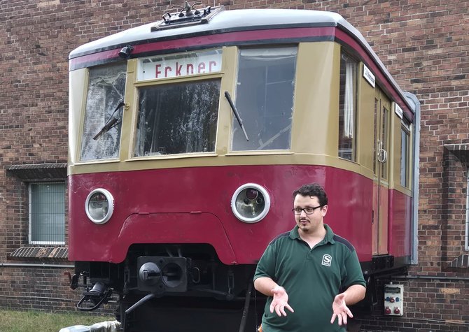 Vorderansicht eines rot-beigen Triebwagens vor einer Backsteinwand. Im Vordergrund steht eine Person in grünem Poloshirt mit aufgedrucktem „S“-Logo. Am Fahrzeug sind zwei runde Scheinwerfer, Scheibenwischer und ein Zielschild mit der Aufschrift „Erkner“ sichtbar.