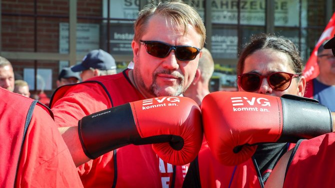 Nahaufnahme von zwei Personen in roten EVG-Westen, die rote und schwarze Boxhandschuhe mit der Aufschrift „EVG kommt an.“ gegeneinander halten. Im Hintergrund sind weitere Personen sowie rote Fahnen vor einem Gebäude mit großen Fenstern zu sehen.