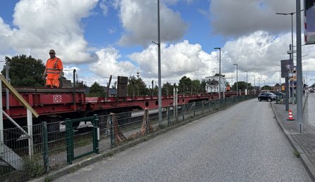 Flachwagen der DB auf einem Gleis neben einer Straße. Eine Person in orangefarbener Warnkleidung steht auf dem Wagen. Im Hintergrund sind Oberleitungen, Masten, Bäume und ein bewölkter Himmel zu sehen.