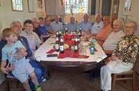 Mehrere Personen unterschiedlichen Alters sitzen an einem länglichen Esstisch in einem Innenraum mit Küchenzeile im Hintergrund. Auf dem Tisch stehen Teller, Gläser, mehrere Flaschen, Brotkörbe und rote Servietten. Eine Person hält ein Kind auf dem Schoß. Die Gruppe blickt in Richtung Kamera.